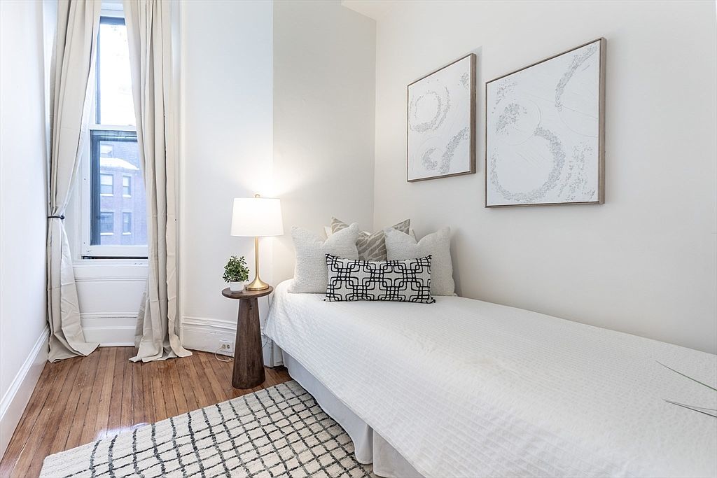 This cozy, minimalist guest bedroom features a single bed dressed in white linens with a decorative patterned accent pillow. A small wooden side table holds a lamp and a potted plant, while two abstract art pieces hang on the wall above the bed. The room is brightened by a tall window draped with neutral curtains and features warm-toned wood flooring covered by a textured area rug.