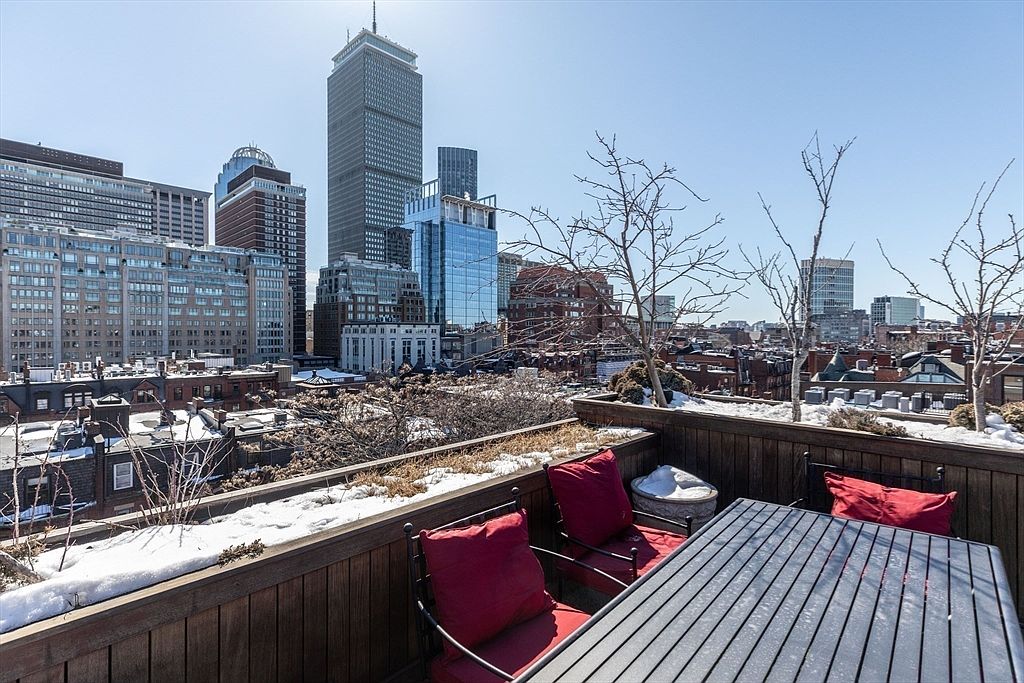 This rooftop deck offers a stunning, elevated perspective of the city skyline, featuring a metal dining table with red-cushioned chairs. The space is bordered by a wooden railing and includes planters with dormant trees and patches of lingering snow, suggesting a crisp, early spring day. The urban backdrop provides a dramatic and sophisticated setting for outdoor entertaining.