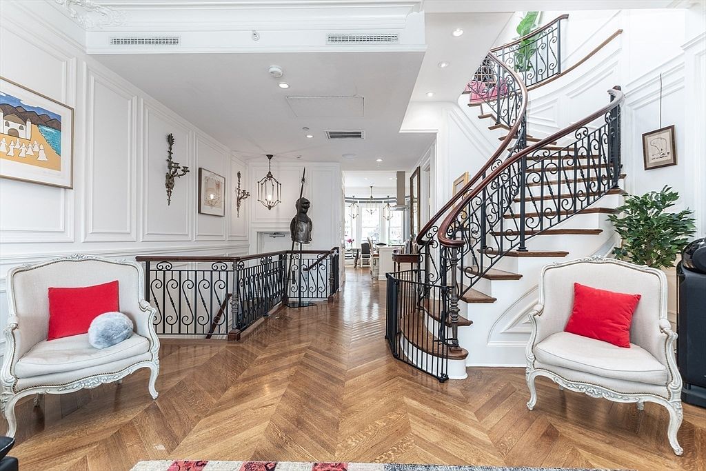 This elegant foyer features a grand curved staircase with intricate wrought-iron railings and polished wood treads, serving as the focal point of the space. The room is finished with classic white wainscoting, herringbone-patterned wood flooring, and two matching armchairs with red accent pillows. A suit of armor stands near the stairwell, adding a unique historical touch to the sophisticated, light-filled interior.