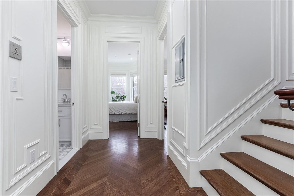 This elegant hallway features rich, dark wood flooring laid in a classic herringbone pattern, complemented by crisp white walls with detailed wainscoting and trim. To the right, a wooden staircase leads upward, while a doorway on the left provides a glimpse into a bright, tiled bathroom. The central passage draws the eye toward a sunlit bedroom at the end of the hall, creating a sense of depth and sophisticated architectural flow.