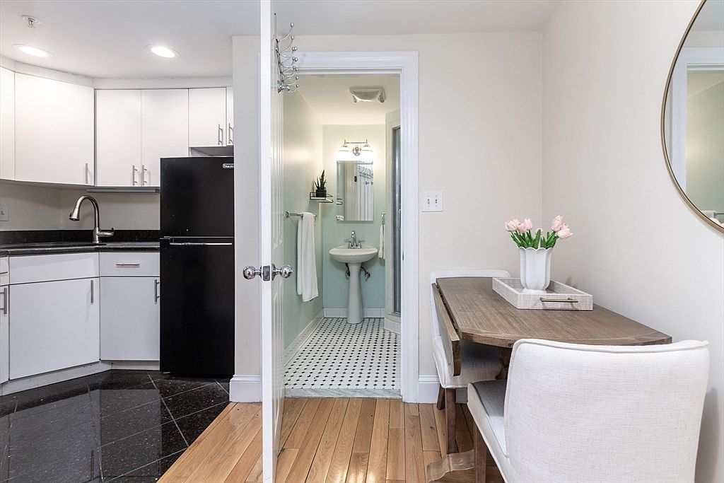 This view captures a compact kitchen area transitioning into a dining space and a bathroom. The kitchen features white cabinetry, a black refrigerator, and dark stone countertops, while the adjacent dining area includes a small wooden table with two upholstered chairs. A doorway leads into a light-toned bathroom with a pedestal sink and patterned tile flooring, creating a functional and efficient layout.