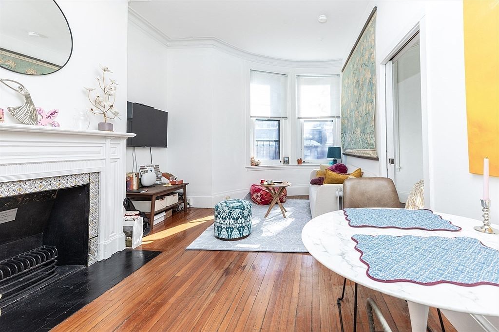 This bright living room features warm wood flooring and a classic fireplace with a white mantel and decorative tile surround. A large bay window allows natural light to fill the space, which is furnished with a comfortable sofa, a patterned ottoman, and a small dining table in the foreground. The room has a cozy, inviting atmosphere with a mix of traditional architectural details and eclectic decor.