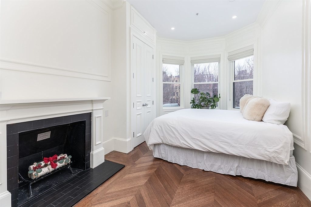 This bright bedroom features elegant herringbone-patterned wood flooring and a classic fireplace with a black tile surround and hearth. A bed is positioned neatly within a bay window alcove, which provides natural light and a view of the neighborhood. The room is finished with crisp white walls, crown molding, and built-in cabinetry, creating a sophisticated and cozy atmosphere.