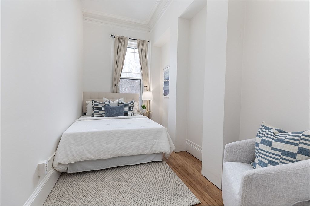 This compact bedroom features a neutral color palette with white walls and light wood flooring. A queen-sized bed with a tufted headboard is centered against the back wall, flanked by a small bedside table and a lamp. To the right, a light-colored armchair with a patterned throw pillow provides a cozy seating area, while a tall window with curtains allows natural light to brighten the space.