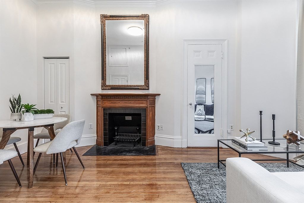 This bright living and dining area features polished hardwood floors and crisp white walls, creating an airy and inviting atmosphere. A prominent wood-mantel fireplace serves as the central focal point, topped by a large, ornate-framed mirror. The space is furnished with a modern dining set on the left and a glass-topped coffee table on the right, offering a balanced layout suitable for both relaxation and entertaining.