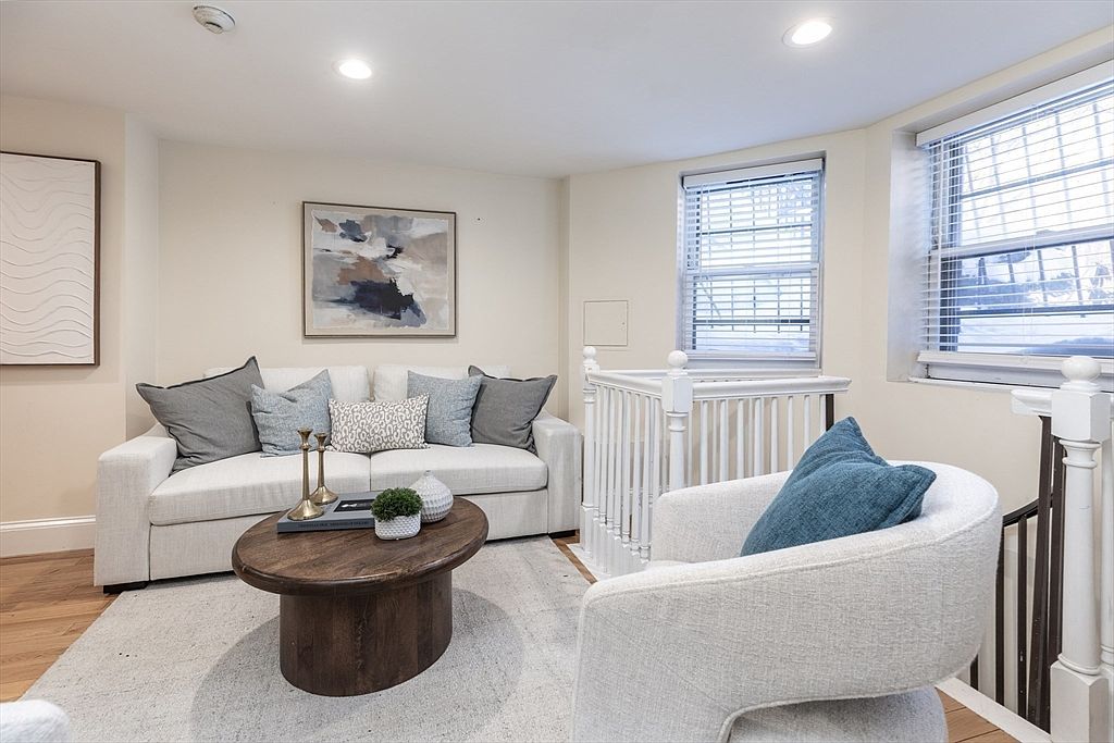 This cozy living room features a light-colored sofa adorned with a mix of gray and patterned throw pillows, centered around a round, dark wood coffee table. A plush, light-toned area rug anchors the seating area, which is complemented by a modern, curved armchair. The space is brightened by natural light from two windows and includes a white wooden railing overlooking a staircase, creating an open and inviting atmosphere.