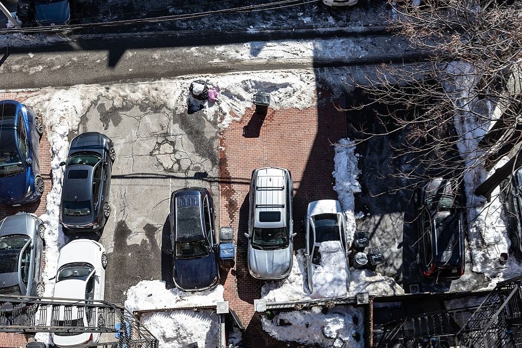 This high-angle aerial perspective captures a residential parking area during winter, featuring a mix of paved and brick surfaces partially covered in snow. Several vehicles are parked in designated spots, with a bare tree extending its branches over the right side of the lot. The scene provides a clear overview of the property's exterior layout and parking capacity.