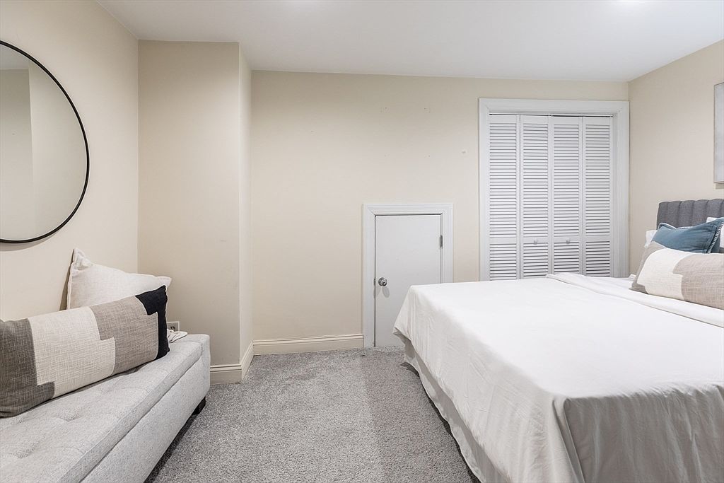 This bedroom features a neutral color palette with soft beige walls and grey carpeting, creating a calm and inviting atmosphere. The room is furnished with a bed dressed in white linens and a long, upholstered bench at the foot of the bed, accented by a patterned throw pillow. A circular mirror hangs on the left wall, while a small, low-profile door and a set of white louvered closet doors add architectural interest to the space.
