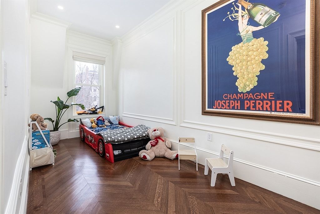 This child's bedroom features rich, dark wood flooring laid in a classic herringbone pattern and crisp white walls with elegant wainscoting. A race-car-themed bed sits near a window, complemented by a large, vintage-style champagne advertisement poster on the wall and two small wooden chairs. The room has a playful yet sophisticated aesthetic, blending classic architectural details with youthful decor.