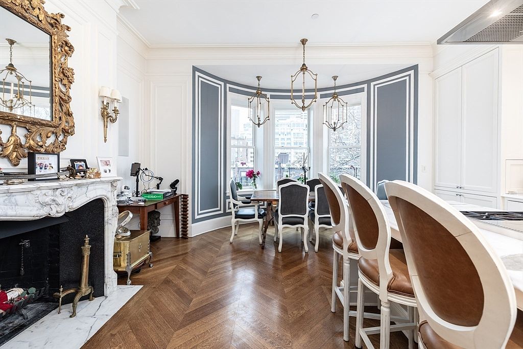 This elegant dining room features a sophisticated blend of classic and contemporary design, highlighted by a prominent bay window area with blue-accented wall molding. The space is anchored by a rich, herringbone-patterned wood floor and a decorative fireplace with an ornate gold-framed mirror above the mantel. A long dining table sits in the bay, complemented by a breakfast bar with high-backed chairs in the foreground, creating a seamless transition between dining and kitchen areas.