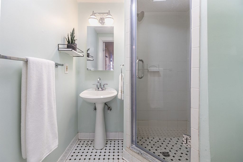This compact bathroom features a classic pedestal sink and a glass-enclosed shower with white wall tiles. The space is accented by a black-and-white patterned tile floor and light-colored walls, creating a clean and bright aesthetic. A small wall-mounted shelf and a towel rack provide functional storage in this efficient layout.