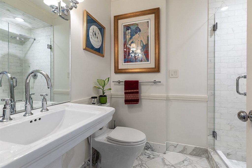 This bright, compact bathroom features a white pedestal sink with polished chrome fixtures and a large wall-mounted mirror. A white toilet sits beside a glass-enclosed shower stall with light-colored patterned tile walls and flooring. The space is accented with framed artwork on the wall and a small potted plant, creating a clean and well-maintained aesthetic.