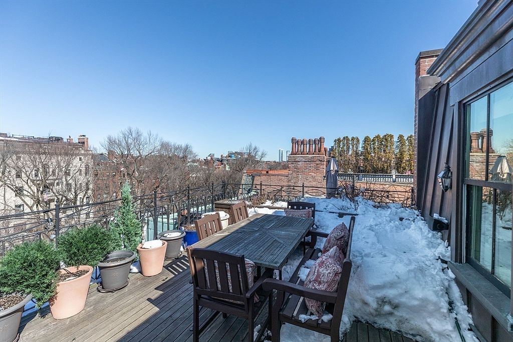 This rooftop deck offers an expansive outdoor living space with a wooden dining set and several potted plants. The area is partially covered in snow, suggesting a winter setting, while a wrought-iron railing provides safety and a clear view of the surrounding urban neighborhood. The perspective captures the deck from an elevated vantage point, highlighting the potential for outdoor entertaining against a backdrop of city architecture.
