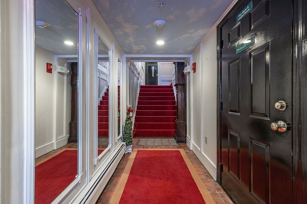This interior hallway features a striking red carpet runner leading to a set of stairs, creating a formal and dramatic entrance. The walls are adorned with large, vertical mirrors that reflect the space, while the ceiling is painted with a decorative sky-and-cloud mural. A black door on the right provides a bold contrast to the light-colored walls, and the overall aesthetic is traditional and grand.
