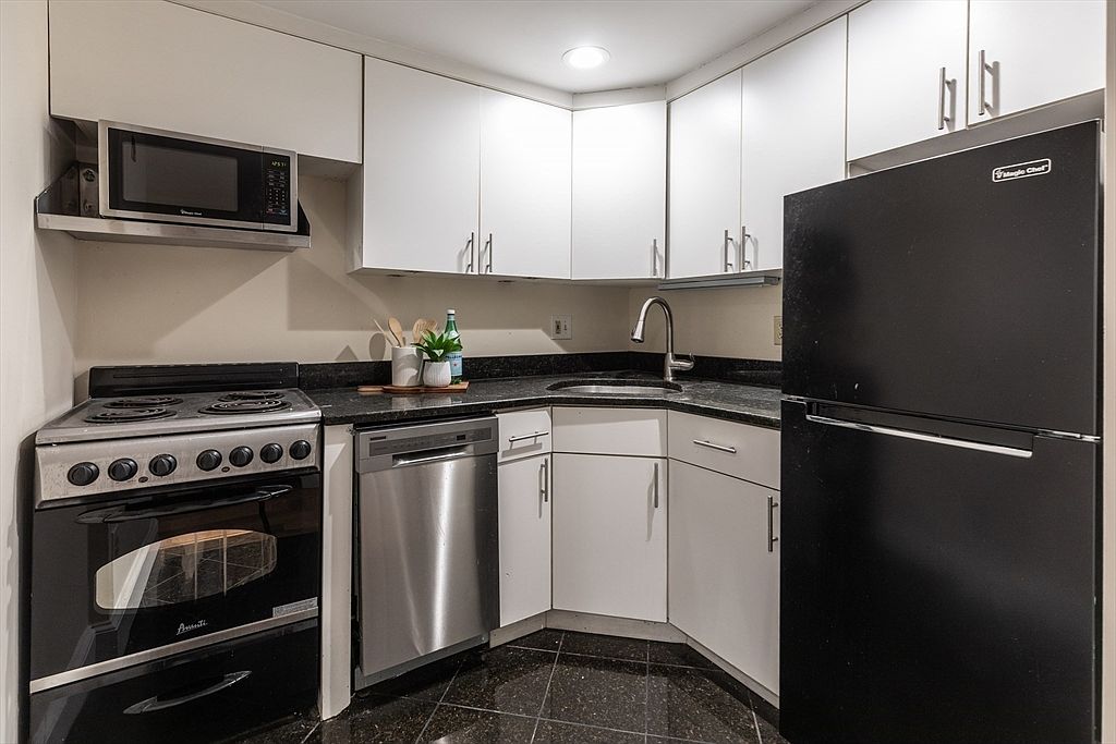 This compact, efficient kitchen features white cabinetry paired with dark stone countertops and dark, speckled flooring. The space is equipped with a stainless steel dishwasher, a black refrigerator, and a black range with an over-the-range microwave. The layout is designed in an L-shape, maximizing the corner area with a stainless steel sink and a gooseneck faucet.