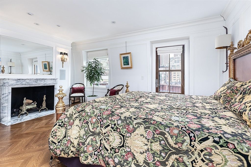 This elegant primary bedroom features a prominent bed with a dark, ornate headboard and a bold, floral-patterned quilt. To the left, a stone fireplace with a decorative mantel serves as a focal point, complemented by a large mirror above it and herringbone-patterned wood flooring throughout. The room is brightened by natural light from a window and a glass-paned door that opens to an outdoor space, creating a sophisticated and classic atmosphere.