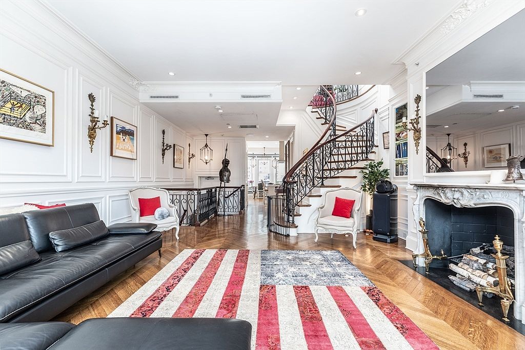 This elegant living room features classic wall molding, rich herringbone wood flooring, and a prominent curved staircase with ornate iron railings. A large area rug with an American flag motif anchors the space, which is furnished with a black leather sofa and two white accent chairs. A traditional fireplace with a decorative mantel serves as a focal point, while the open floor plan leads the eye toward a bright dining area in the background.