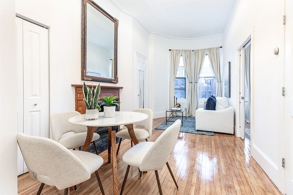 This bright, open-concept dining and living area features warm wood flooring and a neutral color palette. A round dining table with four modern chairs sits in the foreground, positioned in front of a decorative wood mantelpiece topped with a large mirror. Beyond the dining space, a cozy seating area is arranged near a bay window draped with light curtains, creating a welcoming and airy atmosphere.