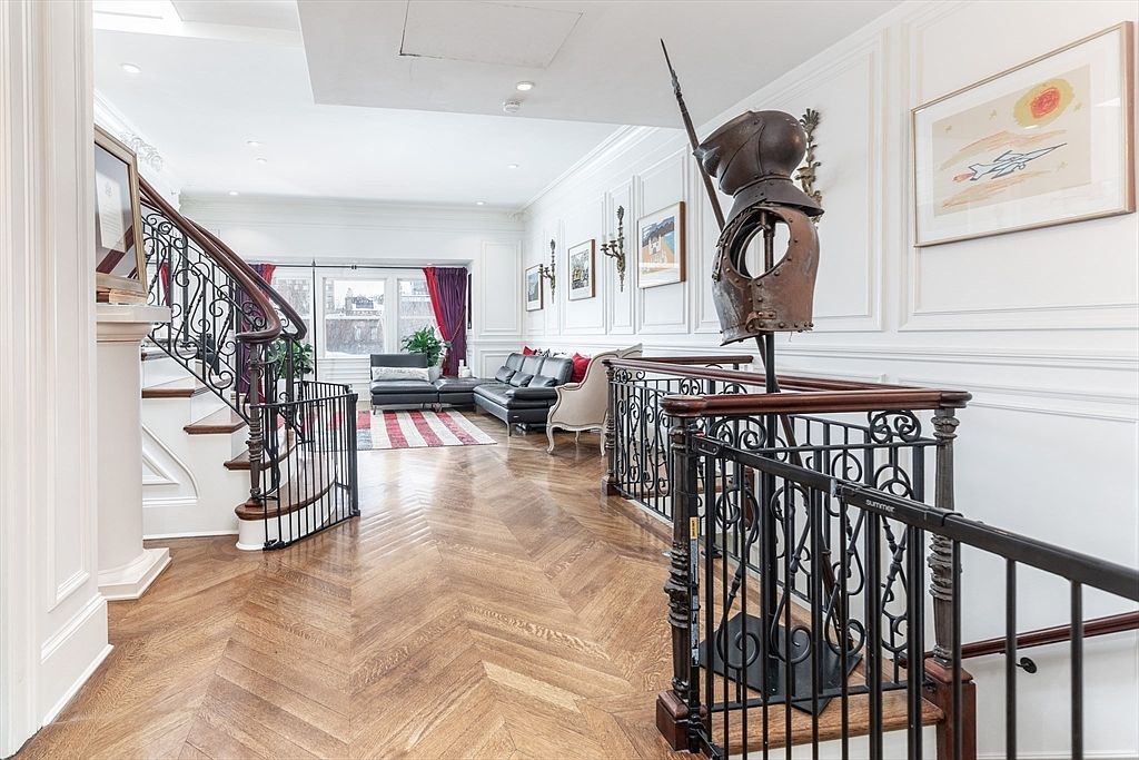 This elegant landing area features rich herringbone-patterned wood flooring and classic white wainscoting along the walls. A prominent suit of armor stands as a decorative focal point near the ornate metal staircase railing, while a bright living area with a black leather sofa is visible in the background. The space is adorned with framed artwork and maintains a sophisticated, traditional aesthetic.