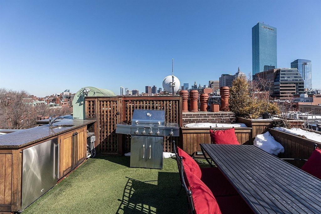 This rooftop deck offers a sophisticated urban outdoor living space, complete with a built-in outdoor kitchen featuring a stainless steel grill and stone countertop. A long dining table with red-cushioned chairs provides ample seating, while the artificial turf flooring adds a touch of greenery against the backdrop of the city skyline. The perspective captures the expansive view of the surrounding architecture, highlighting the property's prime location and potential for entertaining.