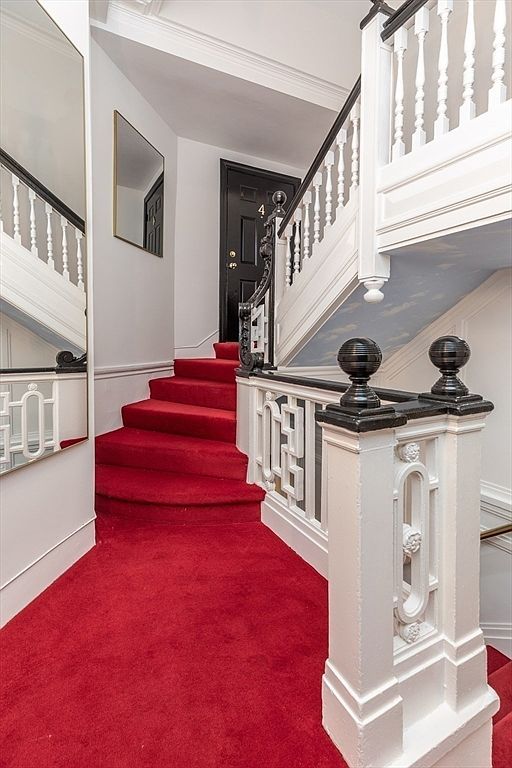 This interior view captures a grand staircase featuring vibrant red carpeting and ornate white balustrades with dark, decorative newel posts. A black door marked with the number 4 stands at the top of the landing, while a large mirror on the left wall reflects the staircase, adding depth to the space. The ceiling above the stairs features a subtle sky-blue painted detail, contributing to a classic and elegant architectural aesthetic.