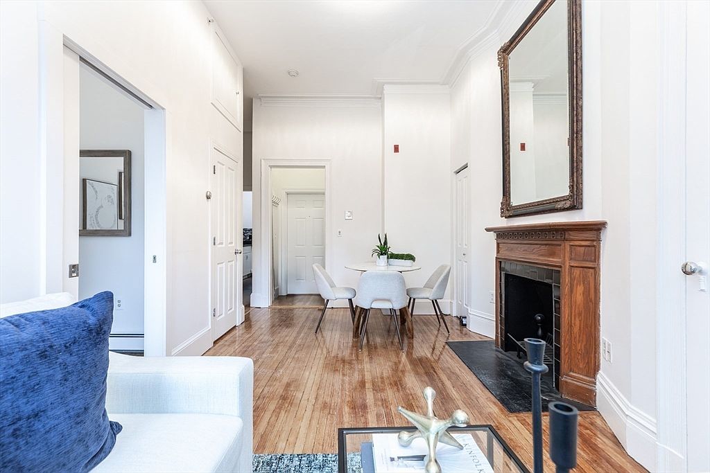 This bright living area features polished light wood flooring and a classic wood-trimmed fireplace mantel topped with a large, ornate mirror. The space flows into a dining area with a round table and three chairs, while white walls and high ceilings create an airy, open atmosphere. The perspective is from the foreground of the living room, looking past a coffee table toward the dining space and a doorway leading to another room.