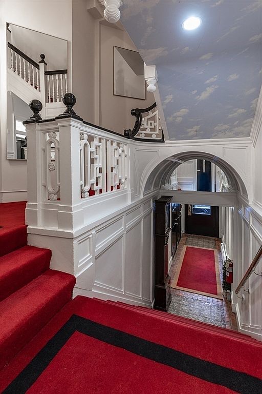 This grand, historic-style entryway features a dramatic staircase covered in a vibrant red runner with a black border. The space is defined by intricate white woodwork, a decorative balustrade, and a unique ceiling painted with a sky-and-cloud mural. A tall, dark wood grandfather clock stands near the arched transition that leads to the front door, creating a sophisticated and welcoming atmosphere.