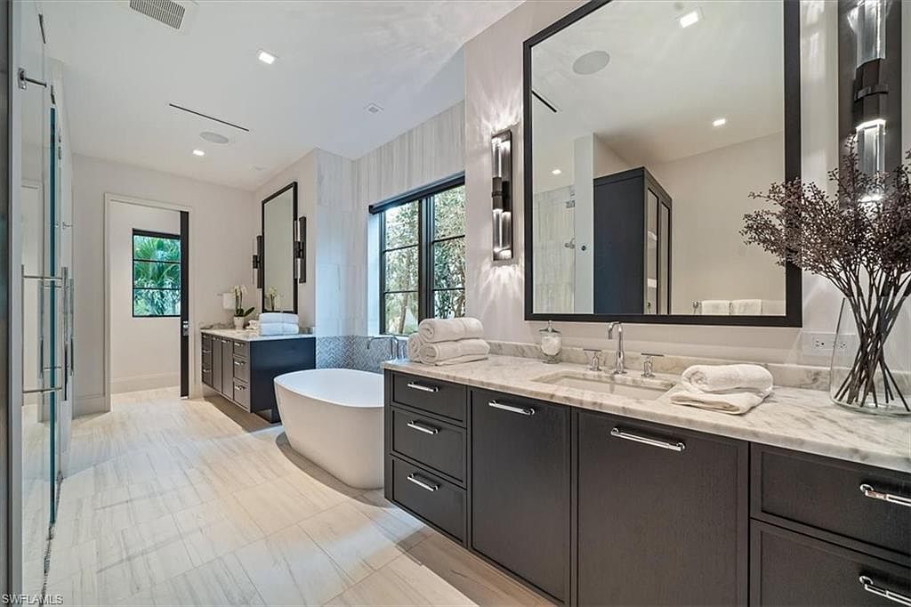 This is a luxurious primary bathroom featuring a freestanding soaking tub, dual vanities with dark cabinetry and marble countertops, and large mirrors with sconce lighting. The space is bright and airy, with light-colored tile flooring and a window providing natural light. The overall impression is modern and spa-like.