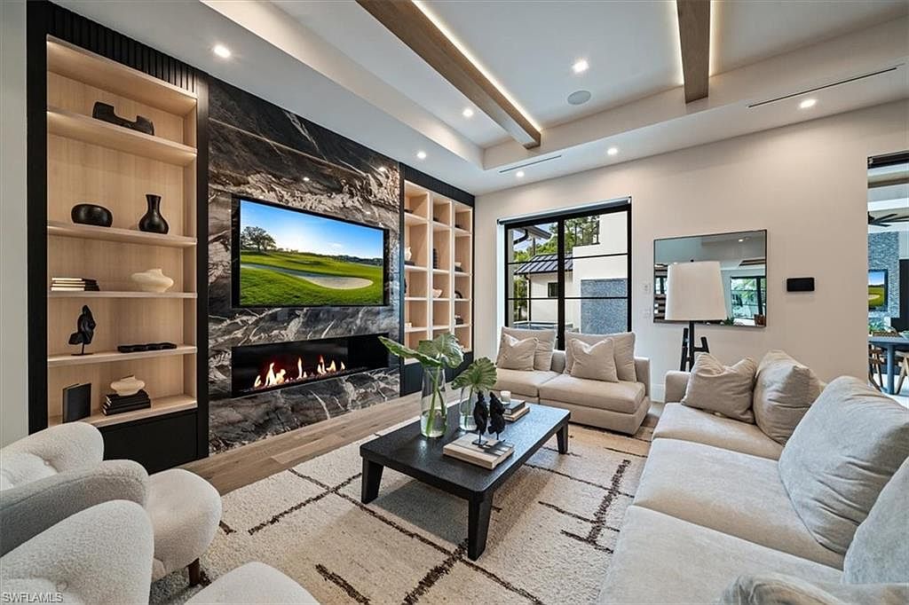 This is an interior shot of a modern living room featuring a fireplace with a marble surround and a built-in entertainment center. The room is furnished with comfortable sofas and a coffee table, and a large window provides natural light. The overall impression is one of luxury and comfort.