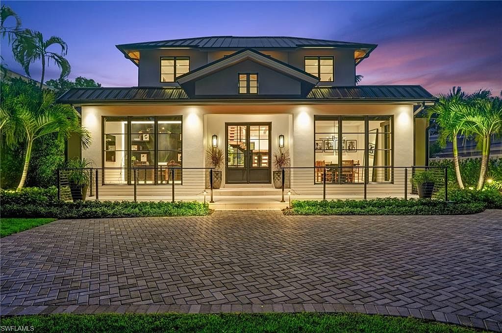 This is a front exterior view of a two-story modern home at dusk. The house features a symmetrical design with large windows, a covered entryway, and a well-manicured front yard with a brick driveway. The lighting highlights the architectural details and landscaping, creating an inviting and luxurious impression.
