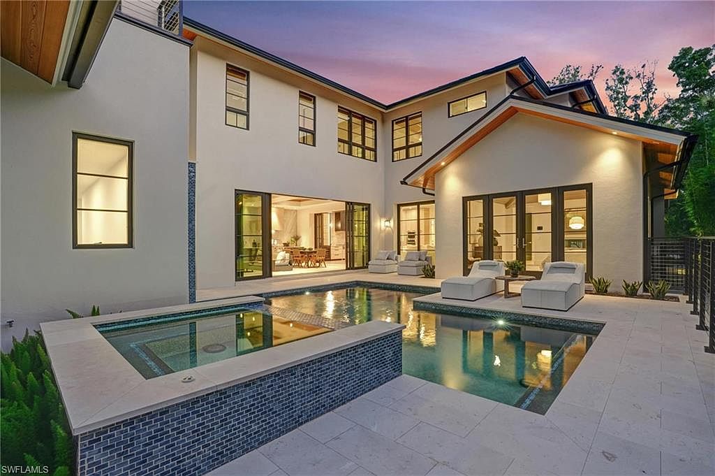 This image showcases the rear exterior of a modern home, highlighting a luxurious pool and spa area. The pool is surrounded by a tiled patio with lounge chairs, and the architecture features large glass doors that open to the interior. The overall impression is one of upscale outdoor living and relaxation.