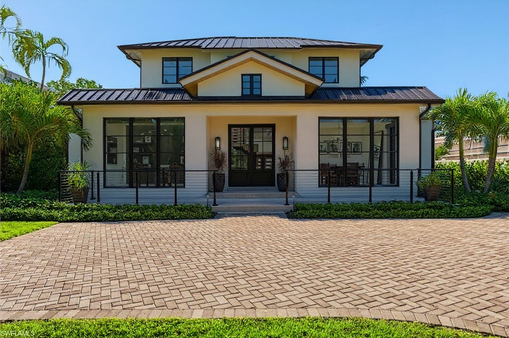 This is a front view of a two-story, light-colored house with a dark metal roof and black framed windows and doors. The exterior features well-manicured landscaping and a brick paver driveway. The home has a modern, elegant design with a symmetry that appeals to the eye.