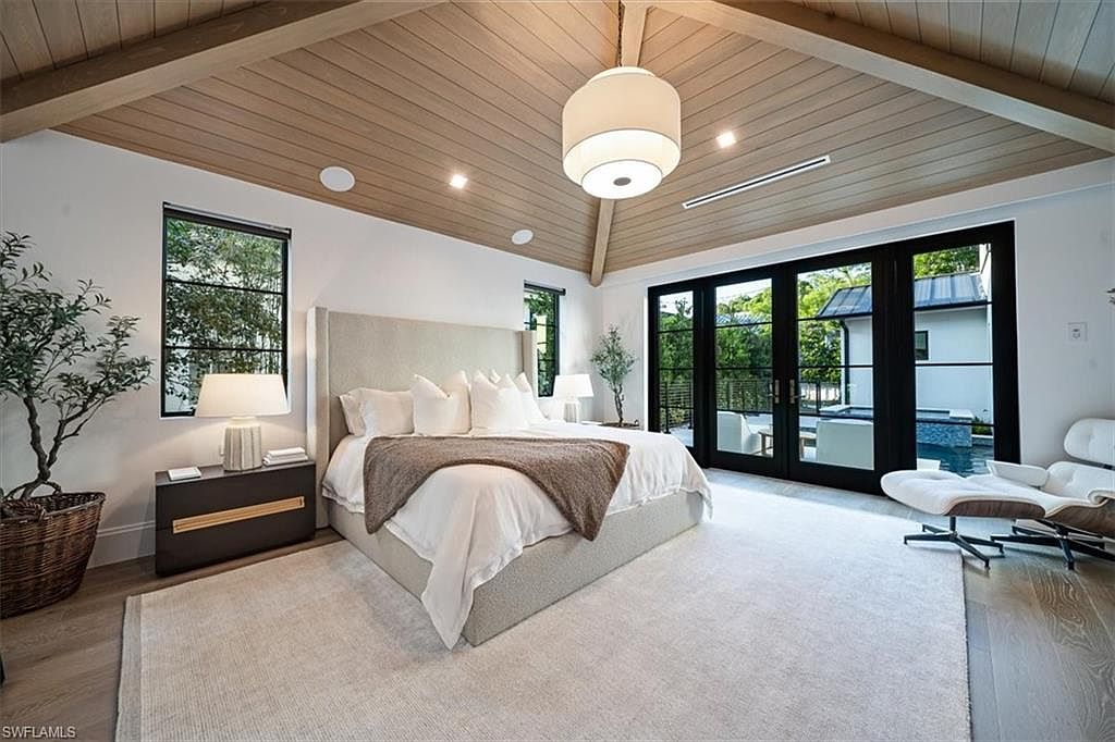 This is a bright and airy primary bedroom featuring a neutral color palette. The room has a large bed with a padded headboard, white linens, and a throw blanket. The ceiling is wood paneled with exposed beams, and there are black framed windows and sliding doors that lead to an outdoor area. The room also includes a modern chair and a side table with a lamp.