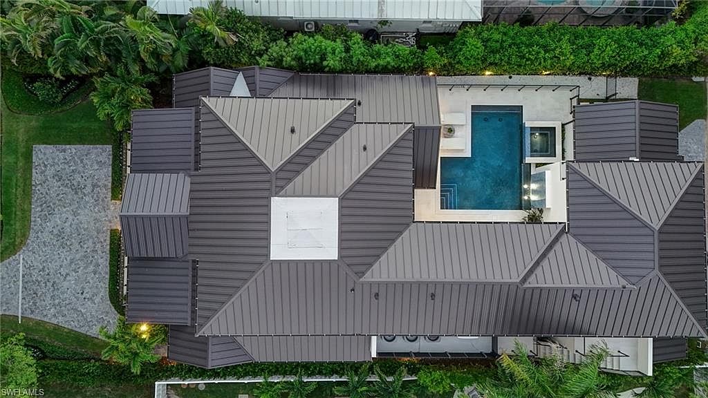 This aerial shot showcases a modern home with a striking dark gray metal roof featuring geometric patterns and a central white section. A rectangular pool and spa are visible in the backyard, surrounded by a light-colored patio. Lush landscaping and mature trees frame the property, adding to its privacy and appeal.