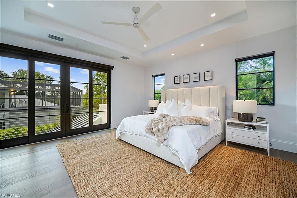 This is a primary bedroom featuring a large bed with a padded headboard, white linens, and a fur throw. The room has a neutral color palette with white walls and a light wood floor, complemented by a large jute rug. Natural light floods the space through large sliding glass doors and a window, enhancing the serene and inviting atmosphere.