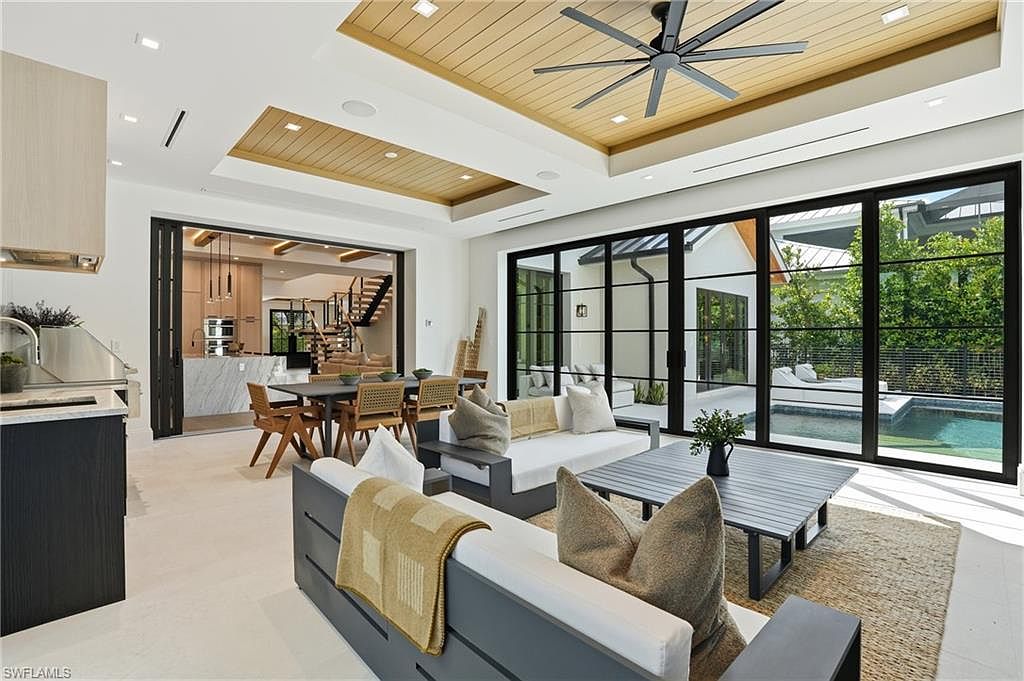 This is an interior shot of a modern living room with a view of the pool area through large sliding glass doors. The room features contemporary furniture, including gray sofas with white cushions and a dark gray coffee table on a jute rug. The ceiling has a unique wood panel design with a modern ceiling fan, and the open floor plan leads to a dining area and kitchen, creating a spacious and inviting atmosphere.
