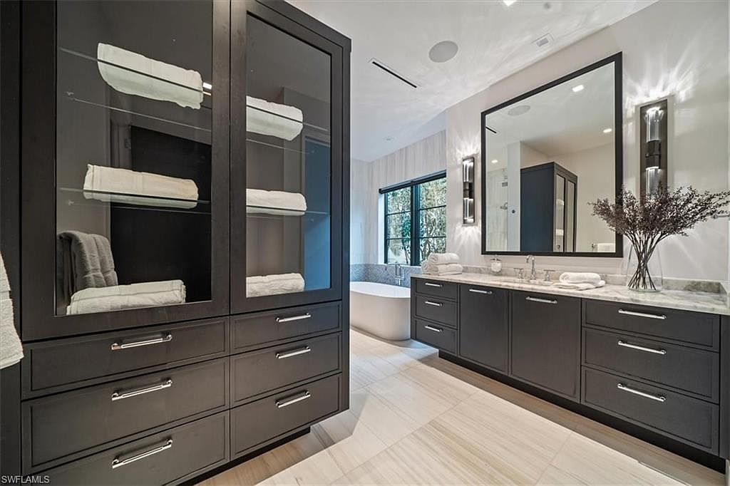 This is a modern primary bathroom featuring dark cabinetry with sleek silver hardware, a large framed mirror, and stylish sconces. A freestanding tub is visible through the window, and the space is well-lit with a clean, contemporary aesthetic. The floor is a light-colored tile, and there is a glass-fronted cabinet for towel storage.