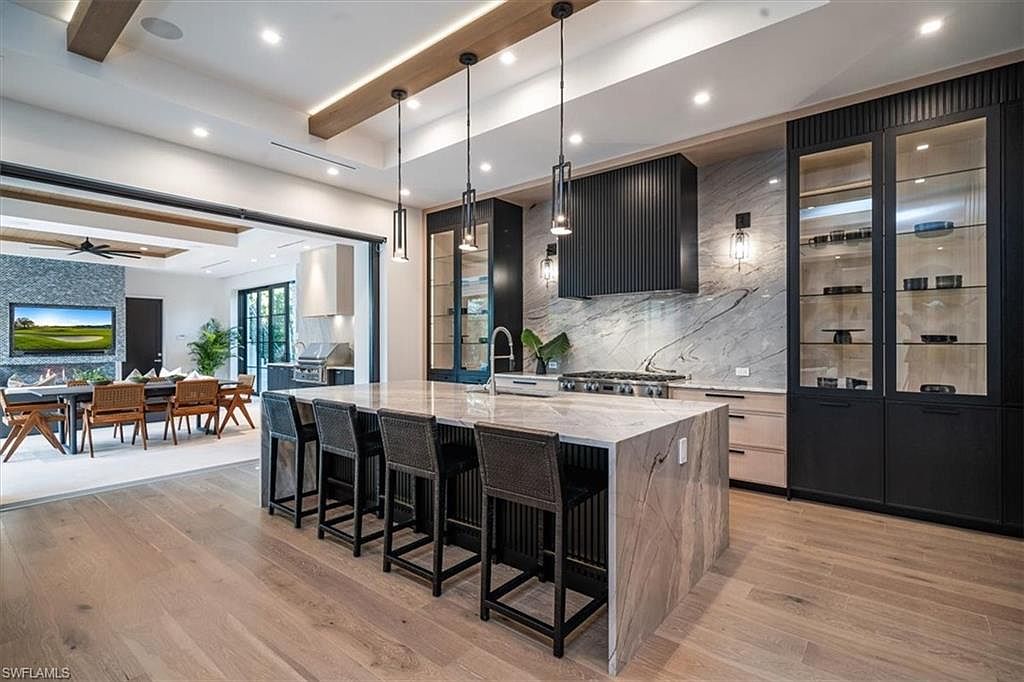 This is a modern, high-end kitchen featuring a large marble island with seating, sleek black cabinetry with glass display cases, and a stylish range hood. The open-concept design flows into a dining area and outdoor space, creating a seamless transition between indoor and outdoor living. The hardwood floors and contemporary lighting fixtures add to the luxurious feel of the space.