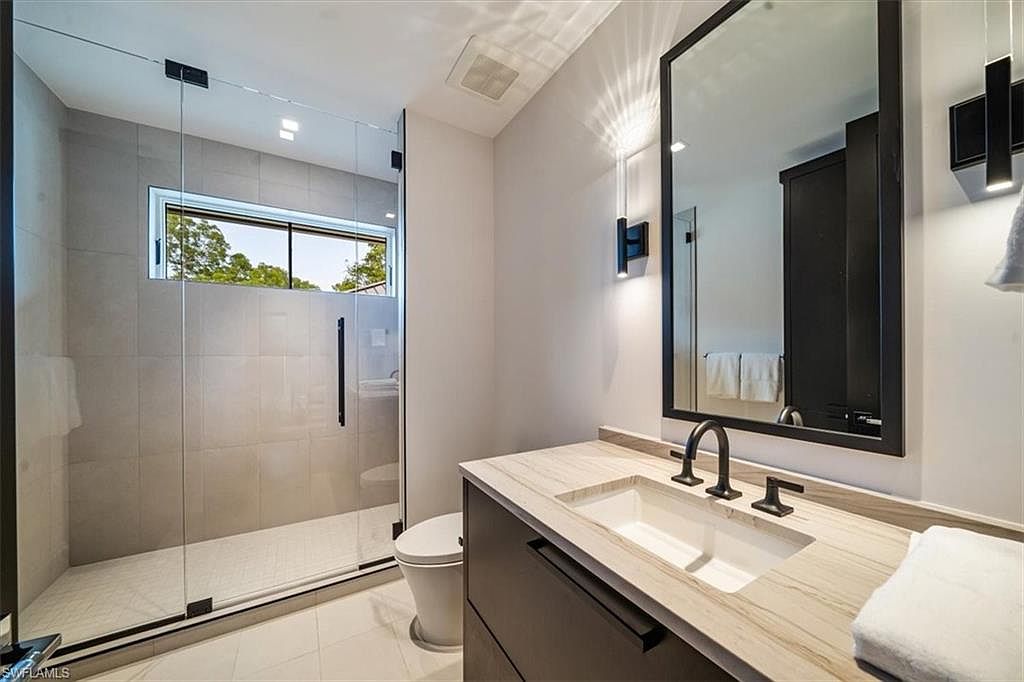 This is a modern bathroom featuring a glass-enclosed shower with a window, a sleek vanity with a light-colored countertop and dark cabinetry, and a black faucet. A large mirror hangs above the vanity, reflecting the clean lines and contemporary style of the space. The overall impression is one of sophistication and functionality.