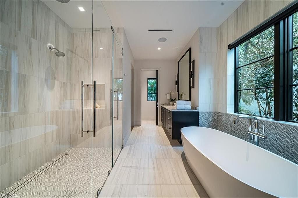 This is a luxurious primary bathroom featuring a glass-enclosed shower with a rainfall showerhead and a linear drain. A freestanding soaking tub sits beneath a large window, complemented by a dark vanity with a marble countertop. The bathroom is finished with light-colored marble-like tile on the walls and floor, creating a spa-like atmosphere.