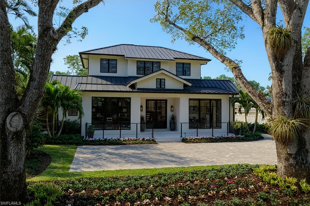 The image showcases the front exterior of a two-story modern home. The house features a white facade, black-framed windows, and a dark metal roof. A neatly landscaped yard with lush greenery and a paved driveway add to the property's curb appeal.