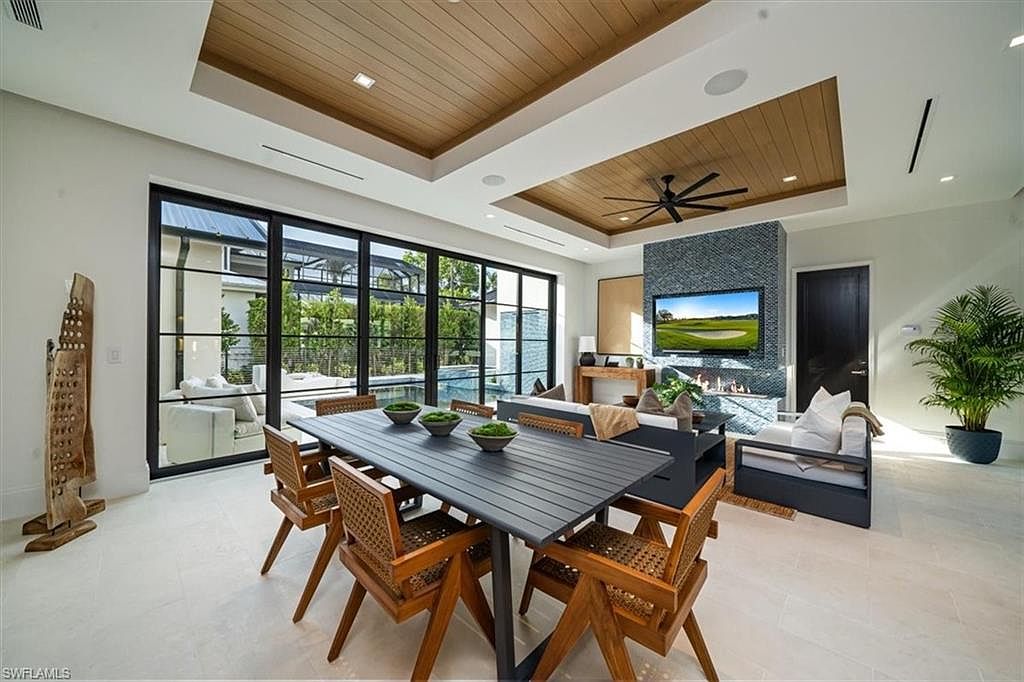This interior shot showcases a modern dining area with a large black dining table surrounded by wooden chairs with woven backs. A wall of windows provides ample natural light and a view of the outdoor pool area. The room also features a living area with a fireplace and television, creating a combined living and dining space with a contemporary design.