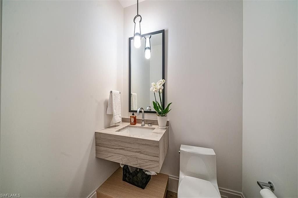 This is a modern guest bathroom featuring a floating vanity with a light-colored stone countertop and a rectangular sink. A black-framed mirror hangs above the sink, illuminated by a pendant light. A white toilet sits to the right of the vanity, and a small orchid adds a touch of elegance to the space. The walls are painted in a neutral color, creating a clean and minimalist aesthetic.