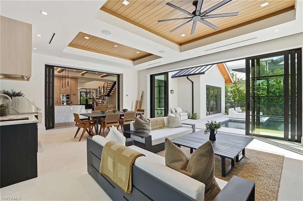 This is an interior shot of a modern living room that seamlessly blends indoor and outdoor spaces. The room features contemporary furniture, including a gray sofa with white cushions and a wooden coffee table, complemented by a neutral-toned rug. Large sliding glass doors open to an outdoor patio with a pool, creating an open and airy atmosphere, while the wooden ceiling adds warmth to the space.