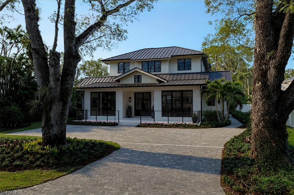 This is a front exterior view of a two-story modern residence. It features a dark metal roof, white exterior walls, and black-framed windows. A brick driveway leads up to the house, and landscaping adds to the curb appeal.