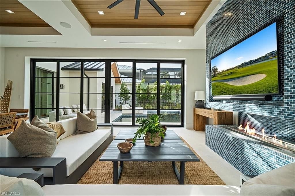 This is a modern living room featuring a large sliding glass door that opens to an outdoor pool area. The room is decorated with a neutral color palette, a sleek sofa, and a contemporary coffee table. A striking blue-tiled wall houses a large television and a modern fireplace, adding a focal point to the space.
