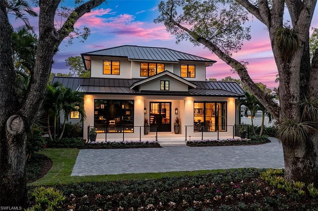 This is a front exterior view of a modern two-story home with a dark metal roof and white facade. The house features large windows, a covered entryway, and a well-manicured front yard with a paved driveway. Mature trees frame the house, adding to its curb appeal and creating a welcoming atmosphere.