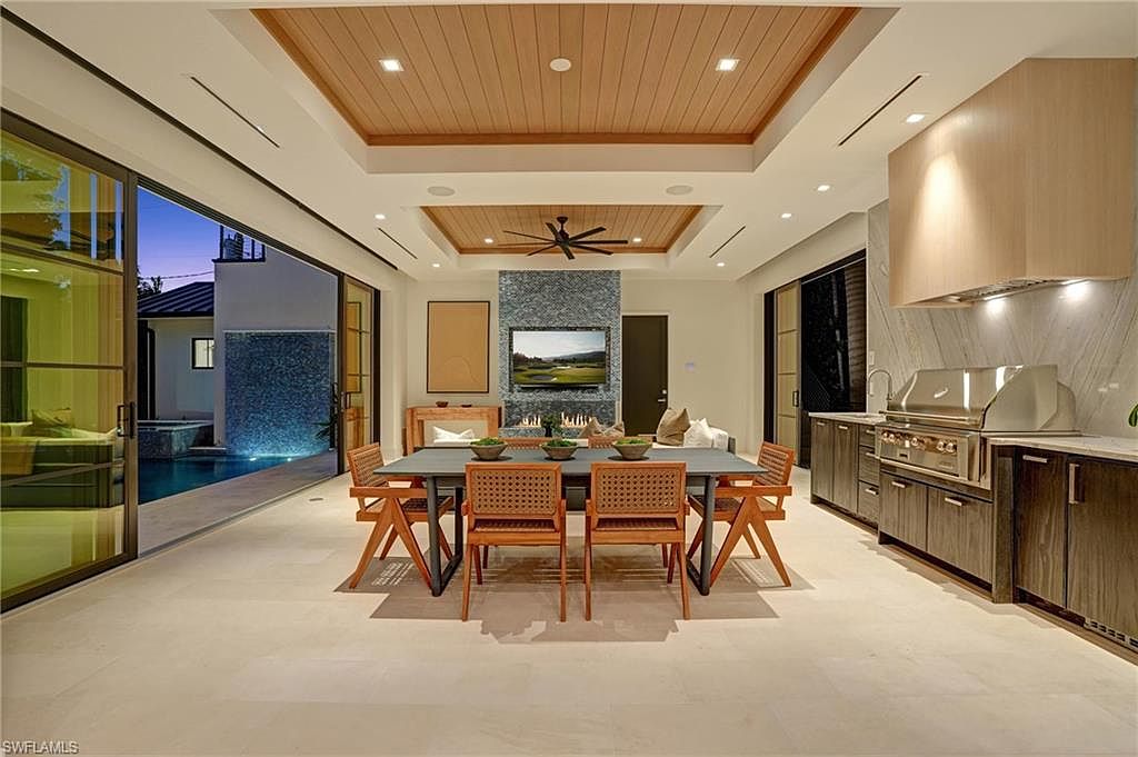 This is an interior shot of a modern dining room that seamlessly blends indoor and outdoor living. The room features a large dining table with seating for eight, a fireplace with a television above, and an outdoor kitchen area. Large sliding glass doors open to a pool area, creating a bright and airy atmosphere.