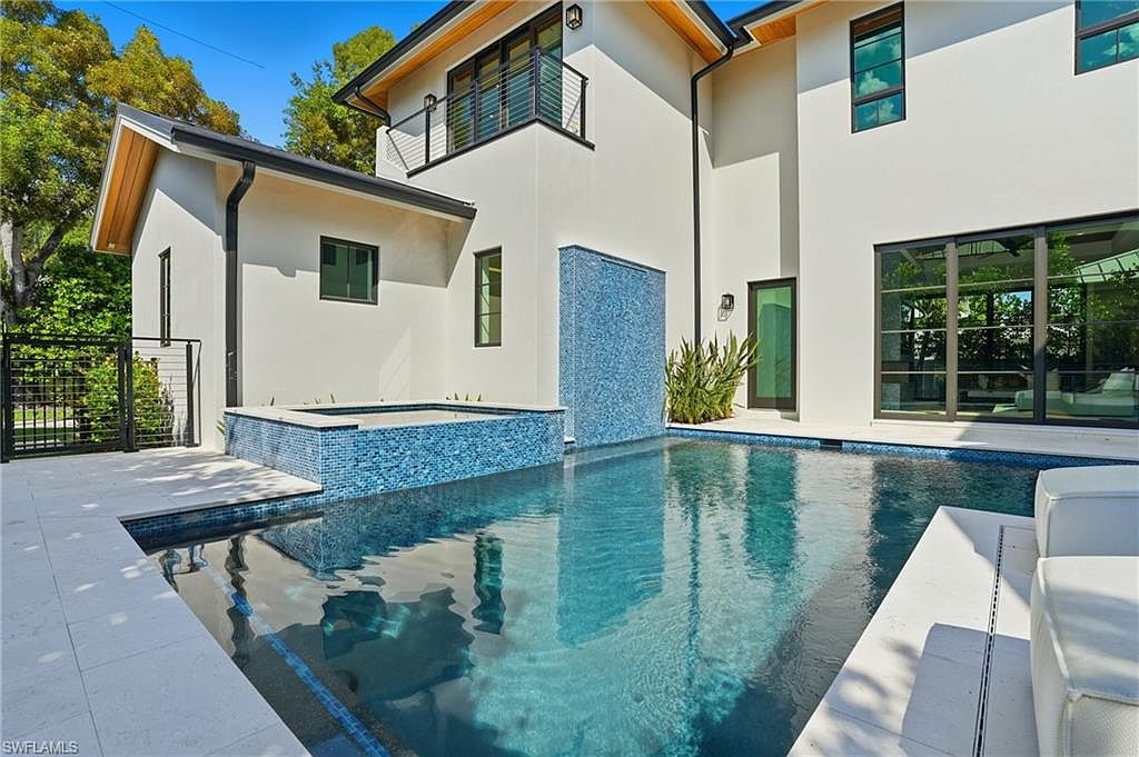 This image showcases a modern home's luxurious pool and spa area. The pool features a unique blue tile design and a waterfall feature, complemented by a built-in spa. The surrounding patio is finished with light-colored stone, and the architecture of the house is sleek and contemporary, with clean lines and large windows that reflect the pool's shimmering water.