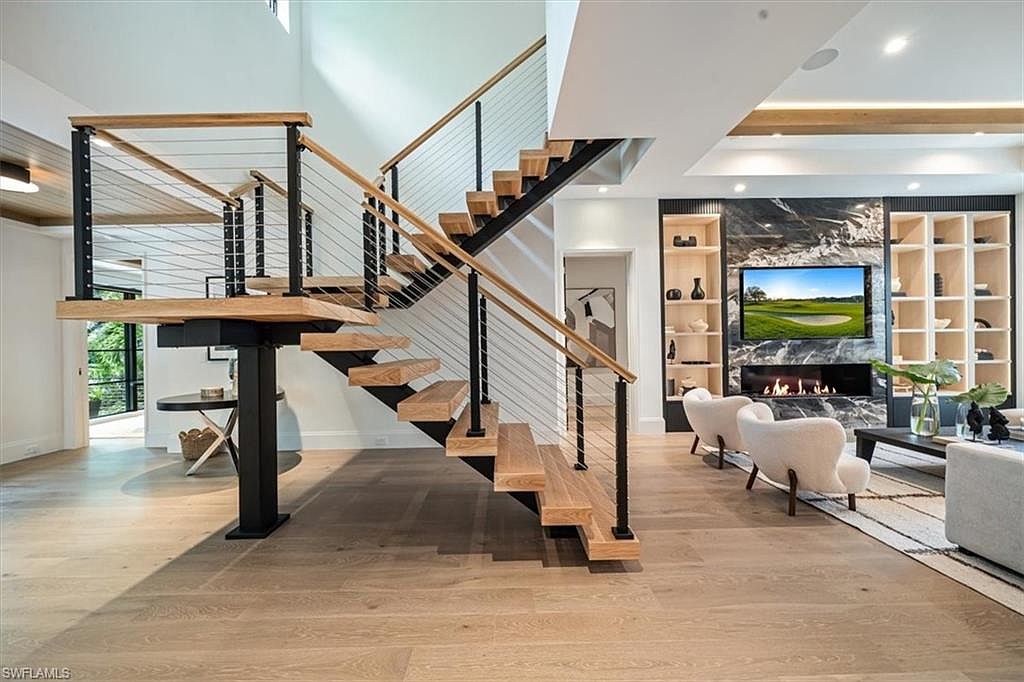 This interior shot showcases a modern living room with a striking staircase as a focal point. The open-concept design features light wood flooring, a built-in entertainment center with a fireplace, and comfortable seating. The staircase has wooden steps with black metal supports and cable railings, adding to the contemporary aesthetic.